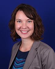 Julie is smiling with shoulder length brown hair and side swept bangs. She is wearing a a blouse with different shades of blue under a gray blazer. She is sitting in front of a dark blue background.