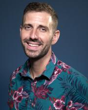 Kellen smilling in front of a blue background wearing a green and magenta floral shirt