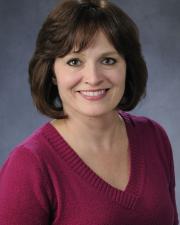 Kim is smiling with shoulder length brown hair and side-swept bangs in a maroon sweater.