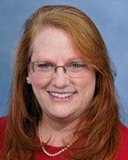 Mary is smiling with long red hair and side-swept bangs wearing a red shirt and pearls.