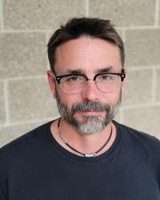 Nate is standing in front of a brick wall, wearing a black t-shirt, black glasses, and a black necklace. He has short brown hair and a mustache and beard.