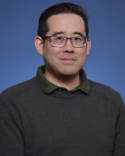 Nicky is smiling against a blue background with a gray sweater, green collar, and wearing glasses