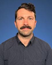Josh sitting in front of a blue background. He's smiling wearing a long sleeve black and white design button down shirt.