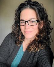 Missy smiling with brown curly hair, black glasses, and a gray sweater over a green shirt